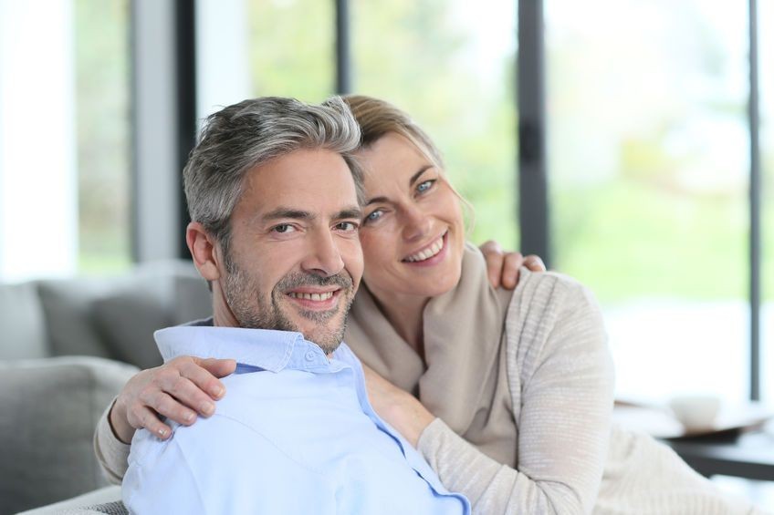 Couple embracing on a sofa in a bright living room with large windows.